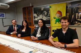 Cuatro personas conversan en una mesa de madera, dos mujeres en chaquetas brillantes y dos hombres con trajes oscuros, en ambiente interior.