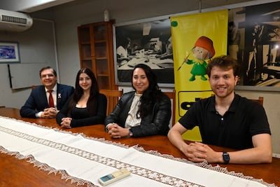 Cuatro personas conversan en una mesa de madera, dos mujeres en chaquetas brillantes y dos hombres con trajes oscuros, en ambiente interior.