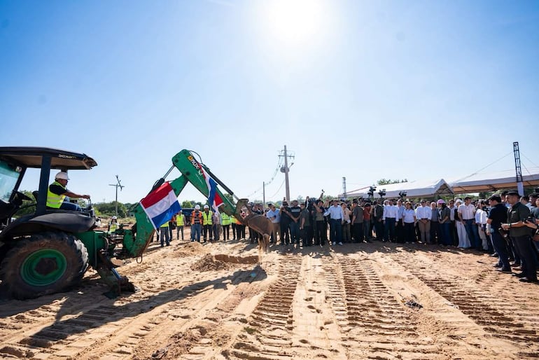Operador en tractor con banderas paraguayas, rodeado de personas con chalecos y cascos en un evento soleado de construcción.