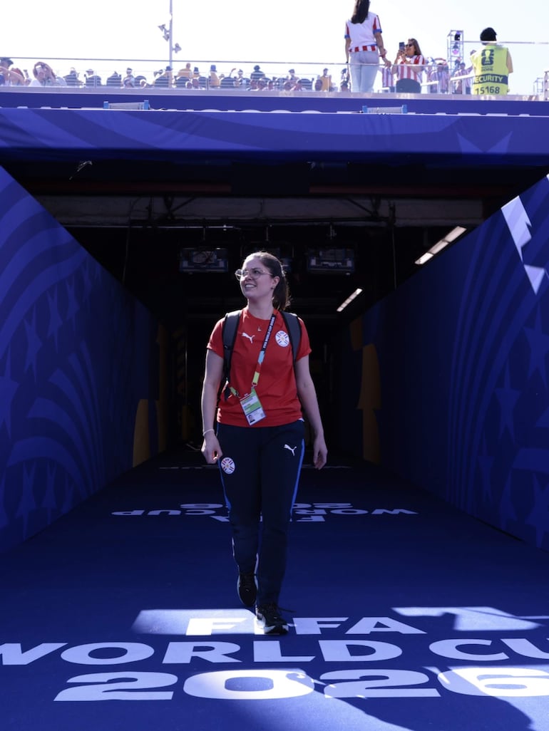 Mujer sonriente en camiseta roja y pantalones Puma, caminando por pasillo hacia la FIFA World Cup 2026, rodeada de gradas llenas.