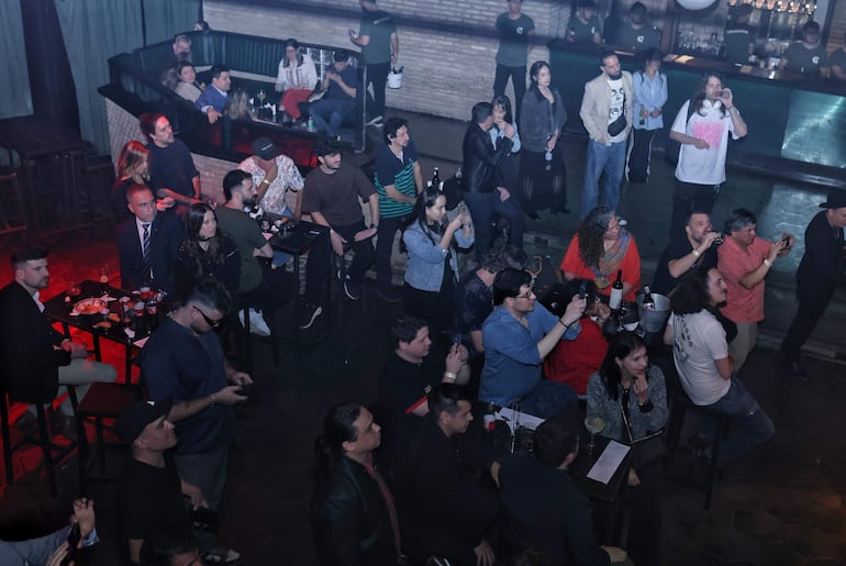 Asistentes en un bar, con luces tenues, conversan y sostienen teléfonos tomando fotos de una actuación en el escenario.