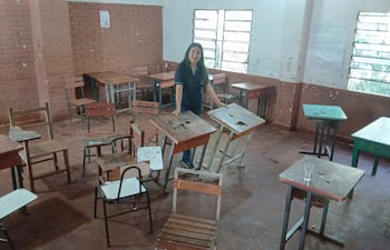 Persona con cabello largo y oscuro organiza escritorios en aula desordenada con sillas y mesas en mal estado.