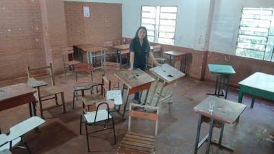 Persona con cabello largo y oscuro organiza escritorios en aula desordenada con sillas y mesas en mal estado.