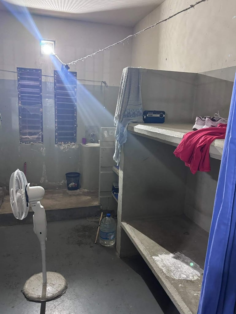 Habitación de prisión con ventilador y radio, ropa colgada y ambiente desordenado.