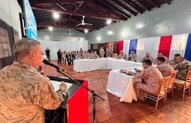 Militar en uniforme habla frente a micrófono, rodeado de miembros de las fuerzas armadas en un salón con colores patrios.