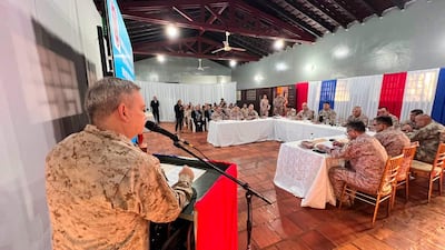 Militar en uniforme habla frente a micrófono, rodeado de miembros de las fuerzas armadas en un salón con colores patrios.
