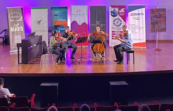 Músicos concentrados, dos en violín, uno en violonchelo y otro en contrabajo, rodeados de luces moradas y partituras.
