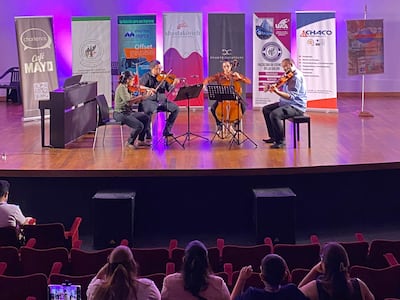 Músicos concentrados, dos en violín, uno en violonchelo y otro en contrabajo, rodeados de luces moradas y partituras.