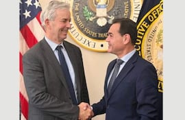 Rubén Ramírez Lezcano y Jeffrey Goettman se estrechan las manos, sonriendo en un entorno oficial con banderas estadounidenses.