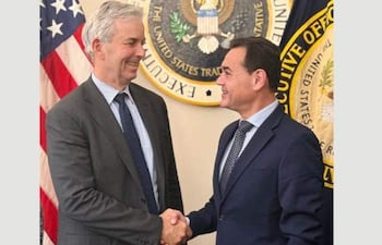 Rubén Ramírez Lezcano y Jeffrey Goettman se estrechan las manos, sonriendo en un entorno oficial con banderas estadounidenses.