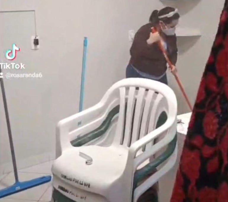 Mujer con mascarilla facial, camisa clara y abrigo marrón utiliza un escobillón en un pabellón con paredes claras.