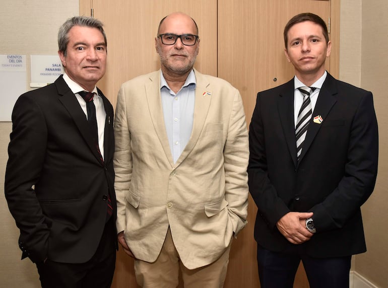 Tres hombres en trajes formales, uno con cabello gris y corbata oscura, otro con chaqueta beige y camisa azul, y un joven con traje oscuro.