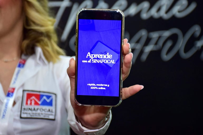 Mujer con camisa blanca y logotipo de SINAFOCAL sostiene un teléfono móvil, mostrando la pantalla. Fondo relacionado con un evento.