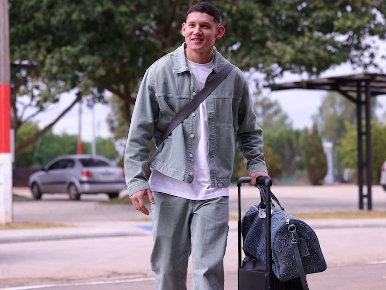 Juan Cáceres sonriente, vistiendo jeans claros y abrigo de mezclilla, lleva un bolso Louis Vuitton y maleta, en un entorno tranquilo.