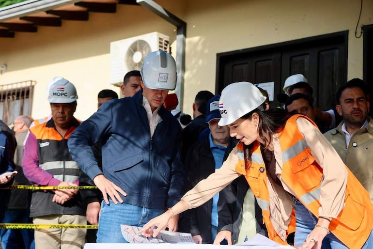 Santiago Peña con abrigo azul y casco observa a Claudia Centurión con chaleco naranja, revisando documentos en una obra pública.