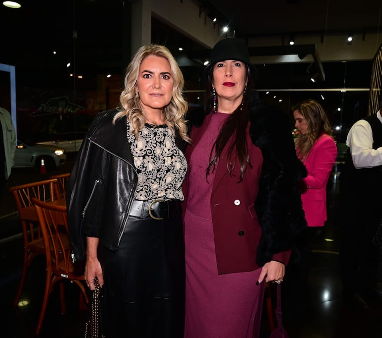Dos mujeres sonríen juntas: una con blusa floral y chaqueta negra, otra en vestido burdeos y chaqueta de pelaje. Fondo moderno.
