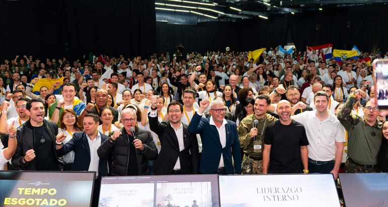 Varias personas levantan los puños en un auditorio, vestidos con camisetas blancas y sosteniendo banderas con colores vivos.
