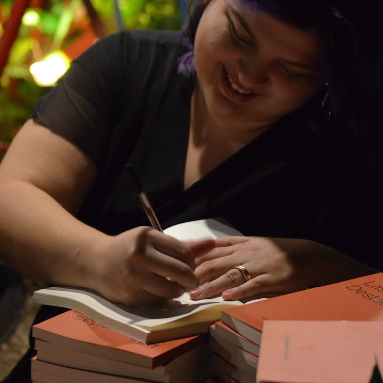 Lía Benítez Flecha, con cabello corto y morado, sonríe mientras firma un libro cubierto de naranja en un ambiente acogedor.
