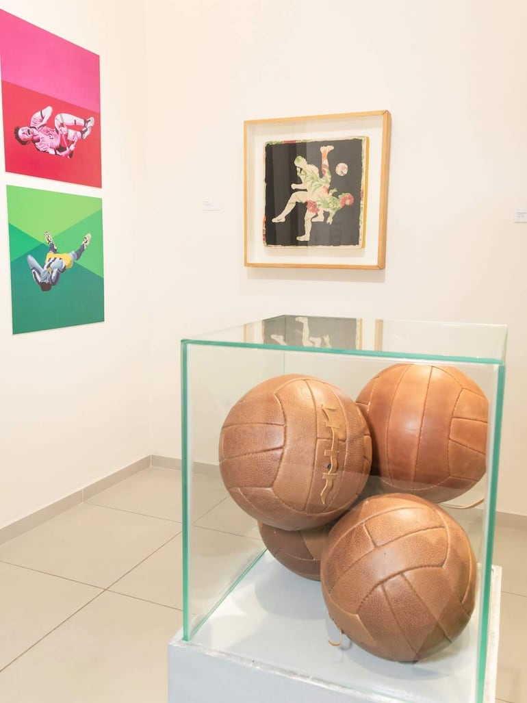 Vitrina con balones de fútbol marrón y tres ilustraciones coloridas de futbolistas en acrobacias en un museo.