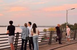 Seis personas conversan en la baranda de un muelle, rodeados de agua y un cielo colorido al atardecer.