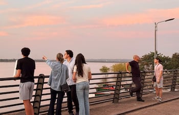 Seis personas conversan en la baranda de un muelle, rodeados de agua y un cielo colorido al atardecer.