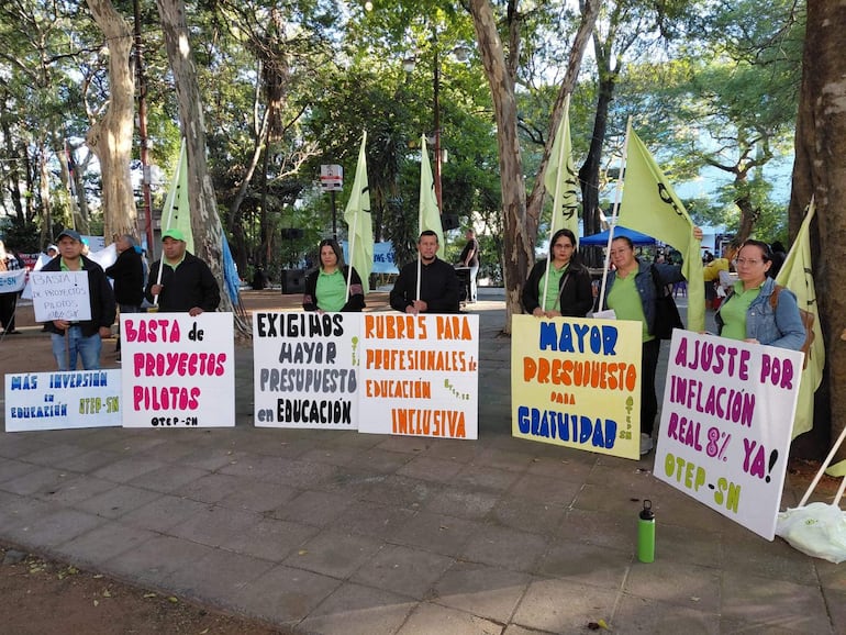 Seis docentes con indumentaria verde sostienen carteles en protesta, rodeados de árboles en un ambiente comunitario.