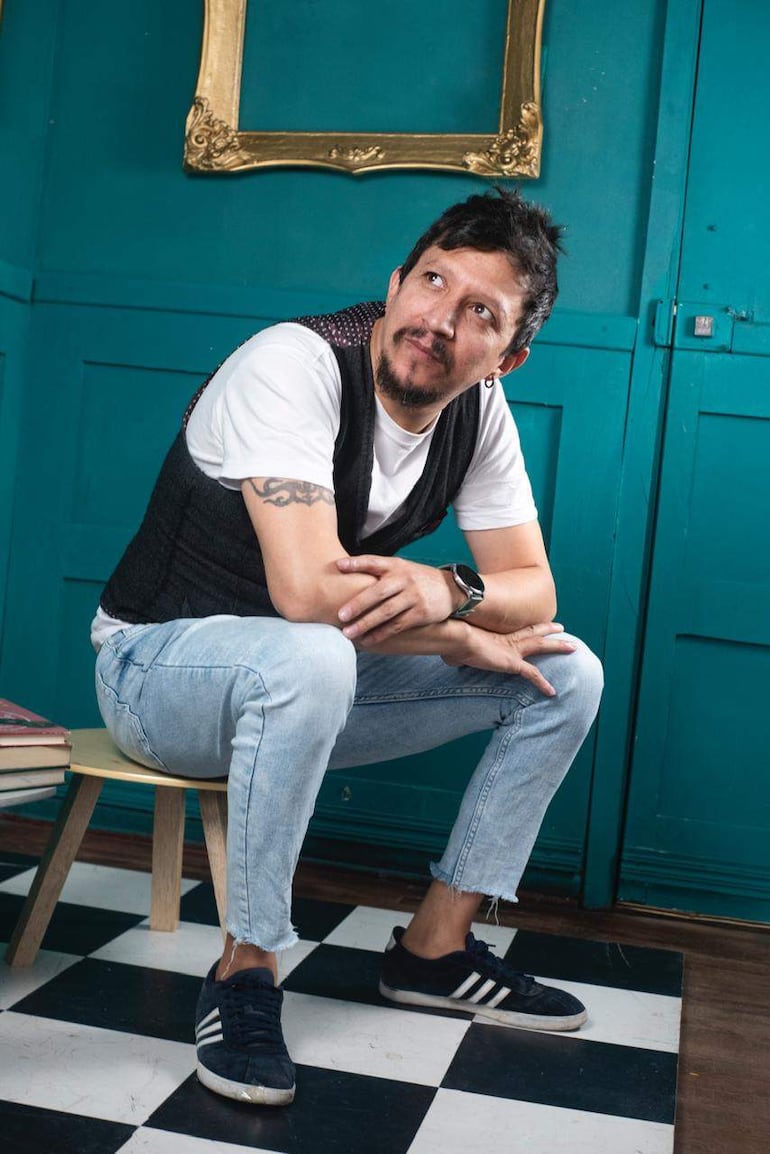 Hombre en camiseta blanca y chaleco negro se sienta pensativo en una silla sobre baldosas en blanco y negro.