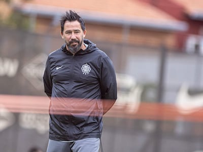 Pablo Sánchez Vitamina en chaqueta negra y pantalones cortos grises, observa a jugadores en un campo de entrenamiento.