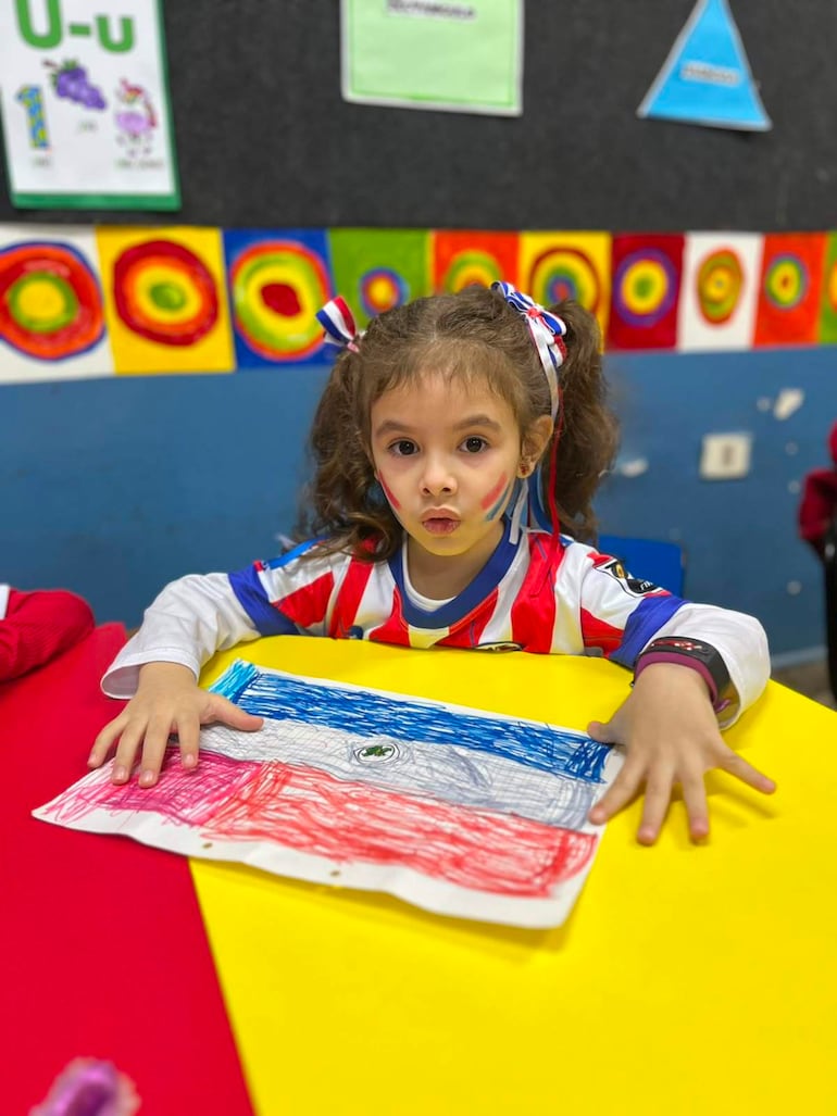 Niña con cabello rizado y cara pintada en rojo, blanco y azul sostiene un dibujo de la bandera, en un salón decorado.