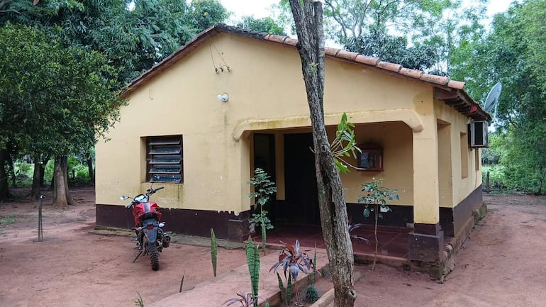 Casa amarilla con base marrón y ventana con rejillas, rodeada de vegetación. Una motocicleta roja está estacionada a la izquierda.