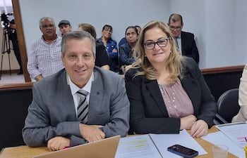 Hombre en traje gris y corbata sonriente junto a mujer rubia en blusa gris, ambos en reunión con documentos y laptops.