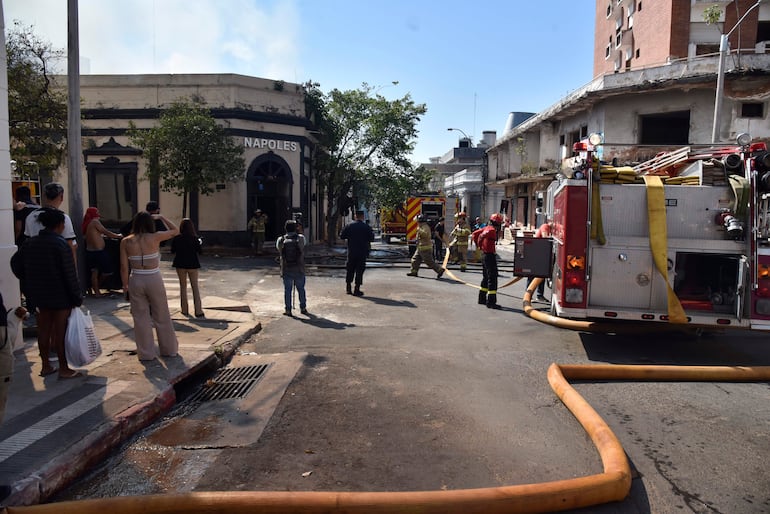 Bomberos en uniformes y cascos, gestionan mangueras y equipos en la calle, mientras espectadores observan la situación desde lejos.