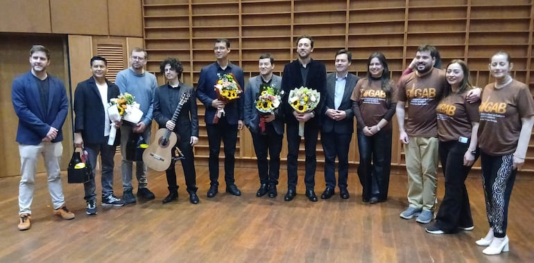 Grupo de diez personas, seis hombres y cuatro mujeres, sonriendo y sosteniendo flores y guitarras en un salón de eventos.