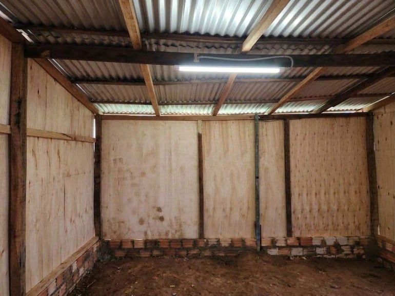 Interior de una estructura rústica con paredes de madera y suelo de tierra, sin mobiliario y con escasa luz natural.