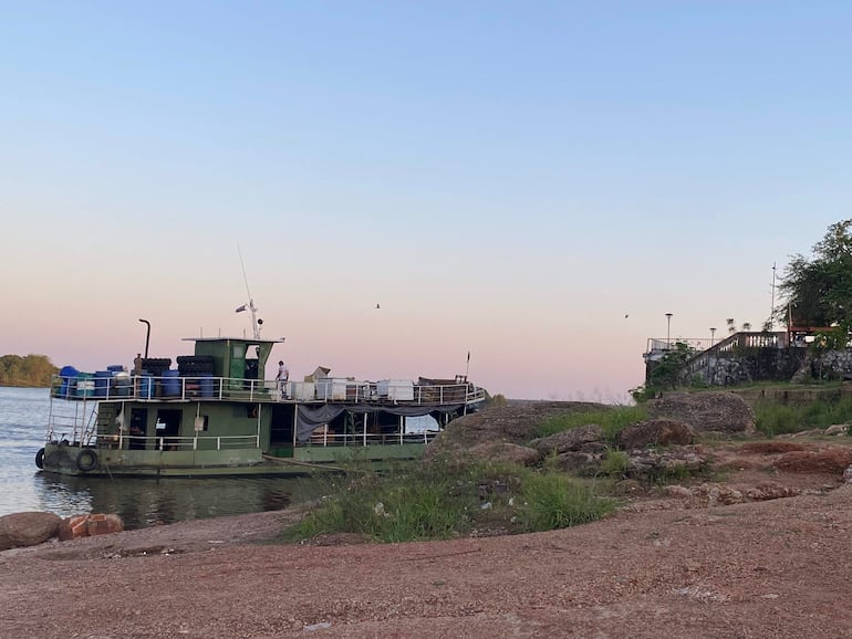 Barco de carga verde atracado, con una persona de pie en la cubierta y objetos visibles en un entorno natural.