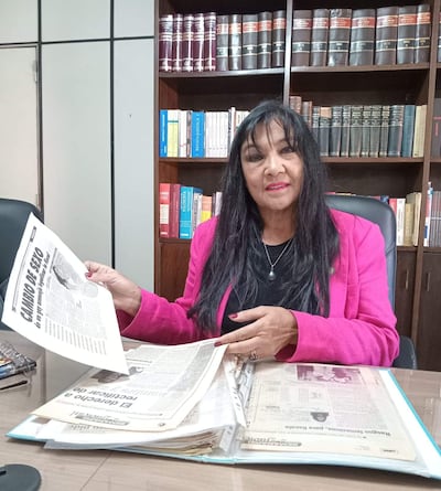 María Gloria Bobadilla, con abrigo rosa, sostiene un periódico mientras revisa documentos en un escritorio lleno de archivos.