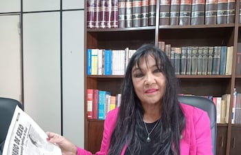 María Gloria Bobadilla, con abrigo rosa, sostiene un periódico mientras revisa documentos en un escritorio lleno de archivos.