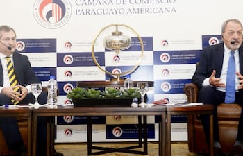 Eugenio Jiménez con traje negro y Víctor Abente Brun con traje oscuro y micrófono, en ambiente formal con logo de la Cámara de Comercio al fondo.