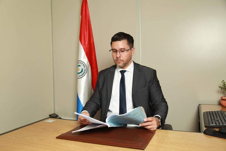 Hombre con traje oscuro y gafas, revisa documentos en una oficina con bandera de Paraguay y planta decorativa.