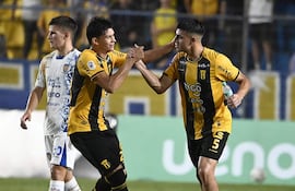 Dos jugadores con camisetas amarillas y negras sonríen y se dan la mano, mientras un tercero en camiseta blanca observa la celebración.