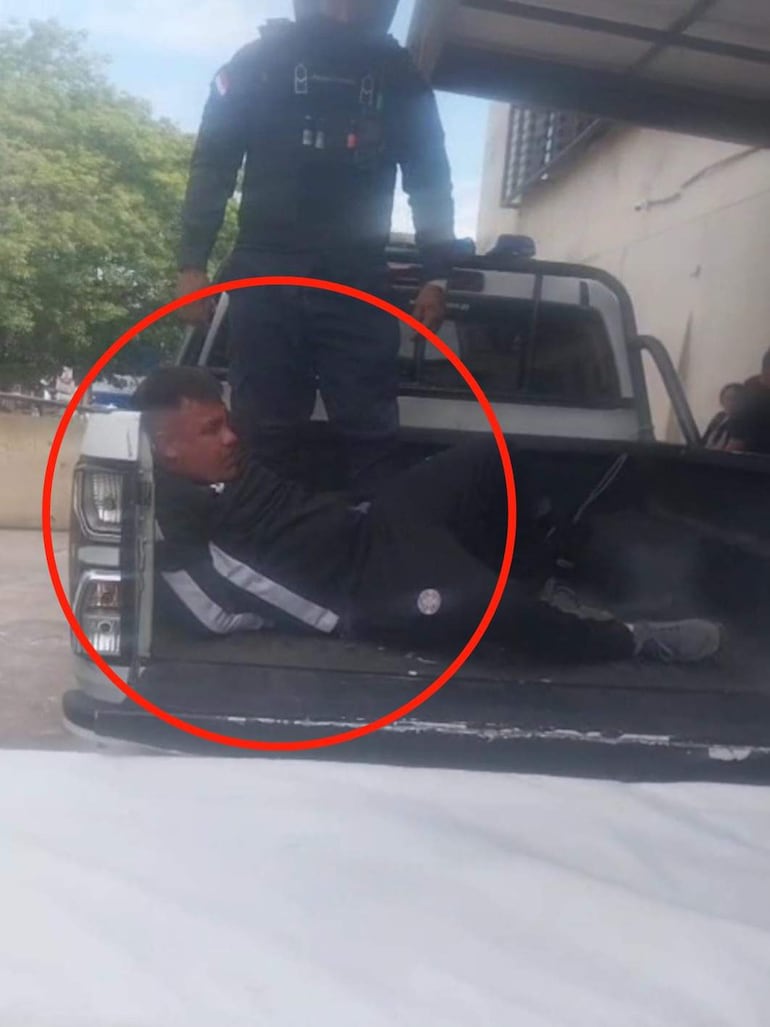 Agente de policía en uniforme negro sobre camioneta junto a hombre reclinado con ropa deportiva oscura.