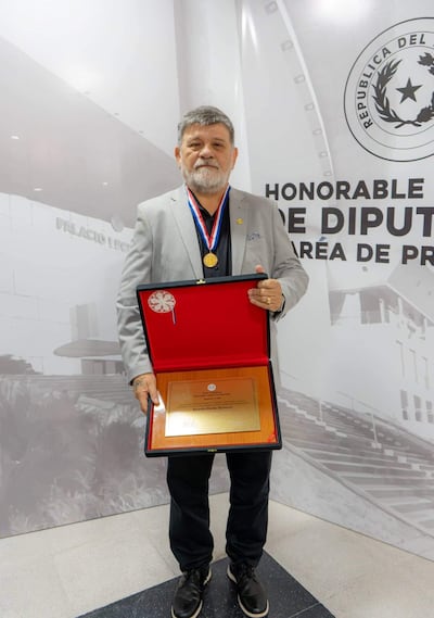 Ricardo Flecha Hermosa, con saco gris y medallón, sostiene una placa en el Palacio Legislativo ante un mural institucional.