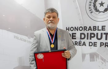 Ricardo Flecha Hermosa, con saco gris y medallón, sostiene una placa en el Palacio Legislativo ante un mural institucional.