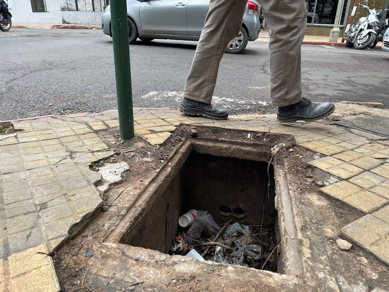 Hombre con pantalones claros y botas negras camina sobre un desagüe abierto repleto de basura en un entorno urbano.