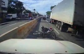 Motociclista con casco en el suelo junto a motocicleta caída en carretera, con vehículos circulando en el área.