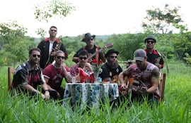 Grupo de hombres en entorno natural. Cuatro sentados serios alrededor de mesa decorada, otros de pie tocando instrumentos.
