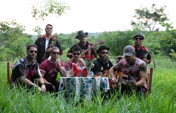 Grupo de hombres en entorno natural. Cuatro sentados serios alrededor de mesa decorada, otros de pie tocando instrumentos.