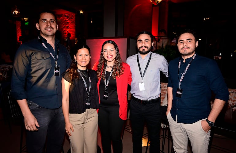 Cinco personas en ambiente elegante, conversando; hombre alto en camisa oscura, mujeres en blusas negras y rojas, hombres con camisas claras y oscuras.