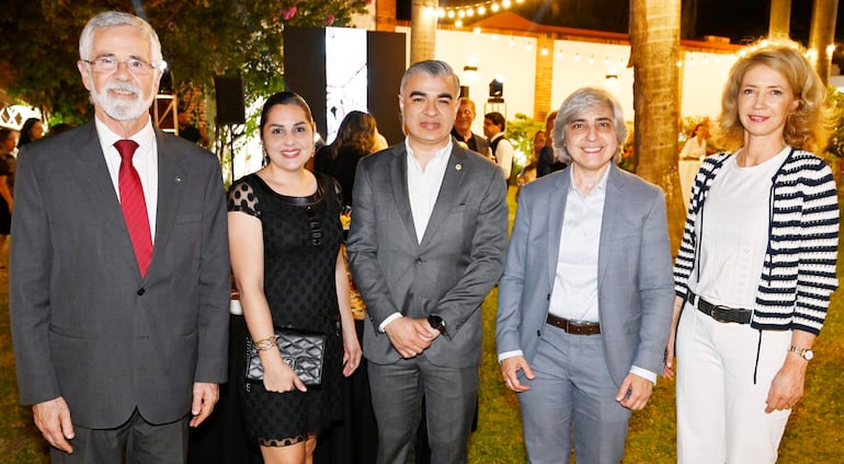 Cinco personas de pie en un evento social, hombre canoso con traje oscuro y corbata roja, mujer con vestido negro y bolso al lado.