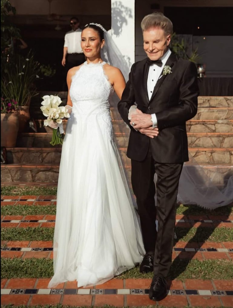 Novia en elegante vestido blanco con ramo de flores y novio con traje negro sonríen en una ceremonia romántica al aire libre.